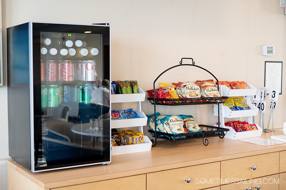 Counter with a small refrigerator on it with drinks inside. Next to it are trays with snacks like granola bars, cookies and chips.