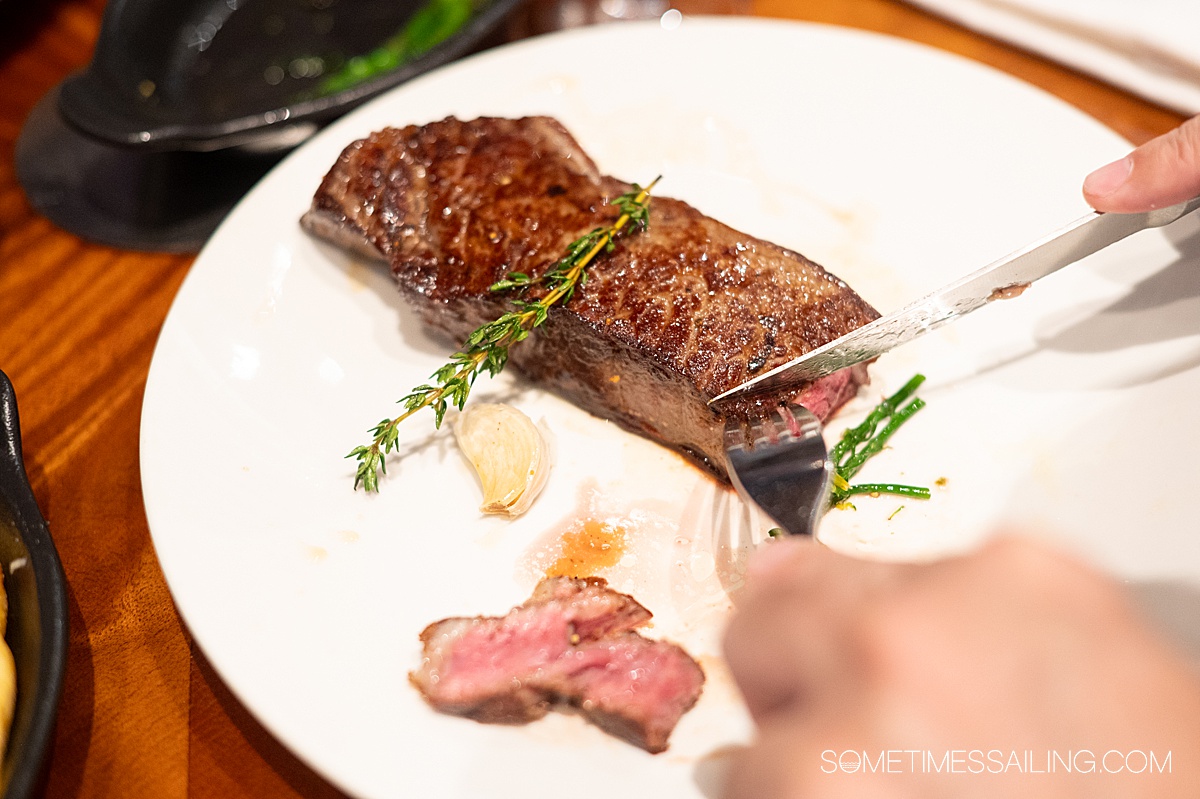 Hands holding a fork and knife, slicing into a steak on a plate at a cruise ship steakhouse.