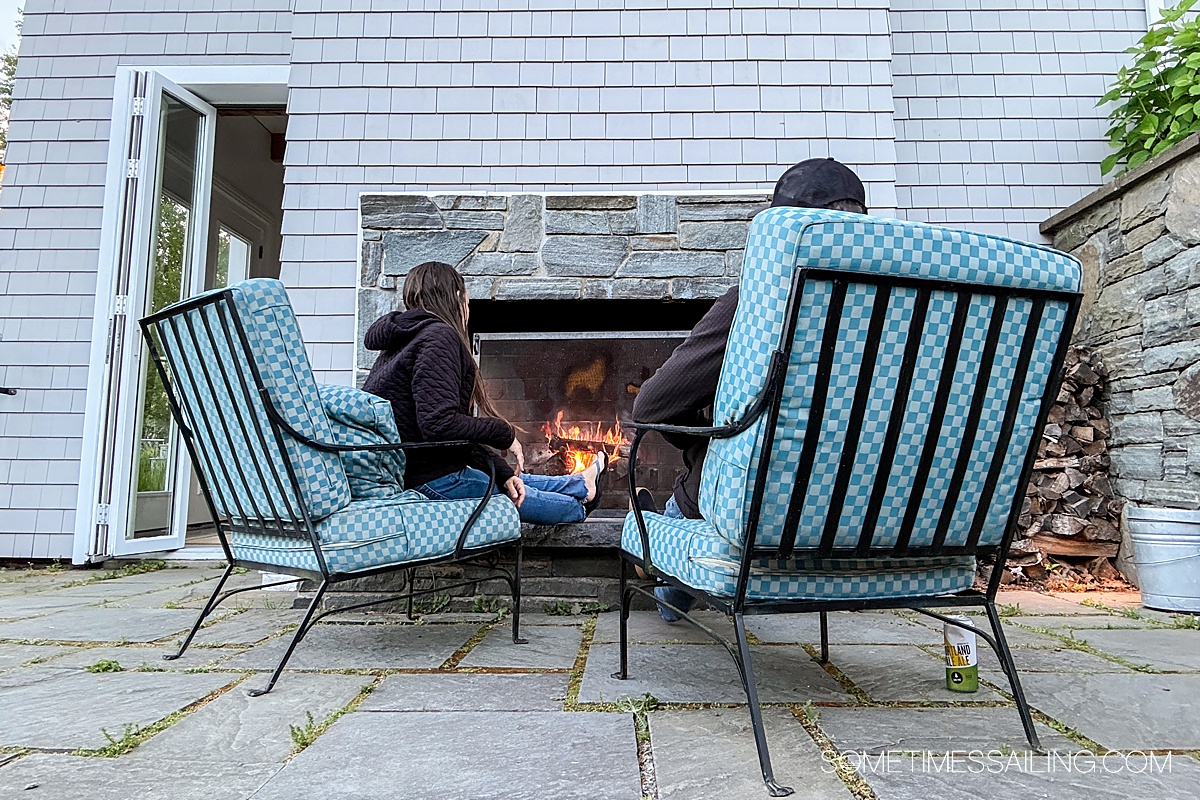 The back of two outdoor chairs with a couple sitting in them looking at an outdoor fireplace.