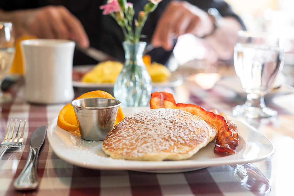 Breakfast plate with a pancake with powdered sugar dusted on top, bacon, and an orange slice.