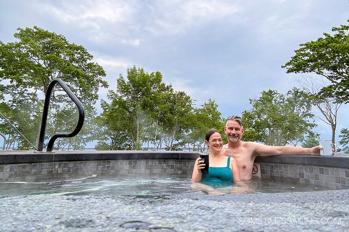 A couple in a hot tub with green trees and a periwinkle sky behind them.
