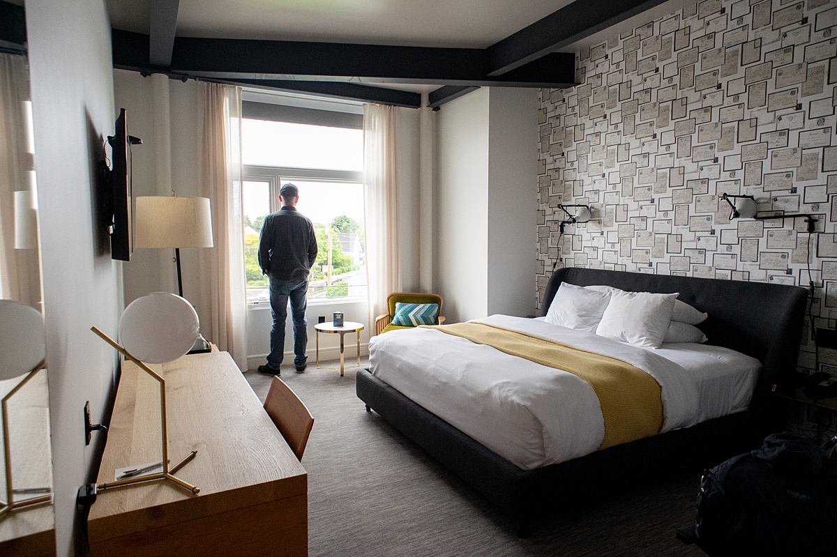 Man looking out the window of a hotel room with a bed and dresser/desk.