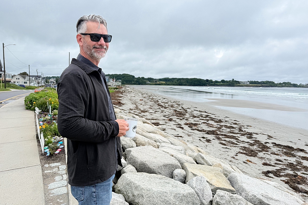 Man with a cup of coffee in his hand and sunglasses on at the beach.