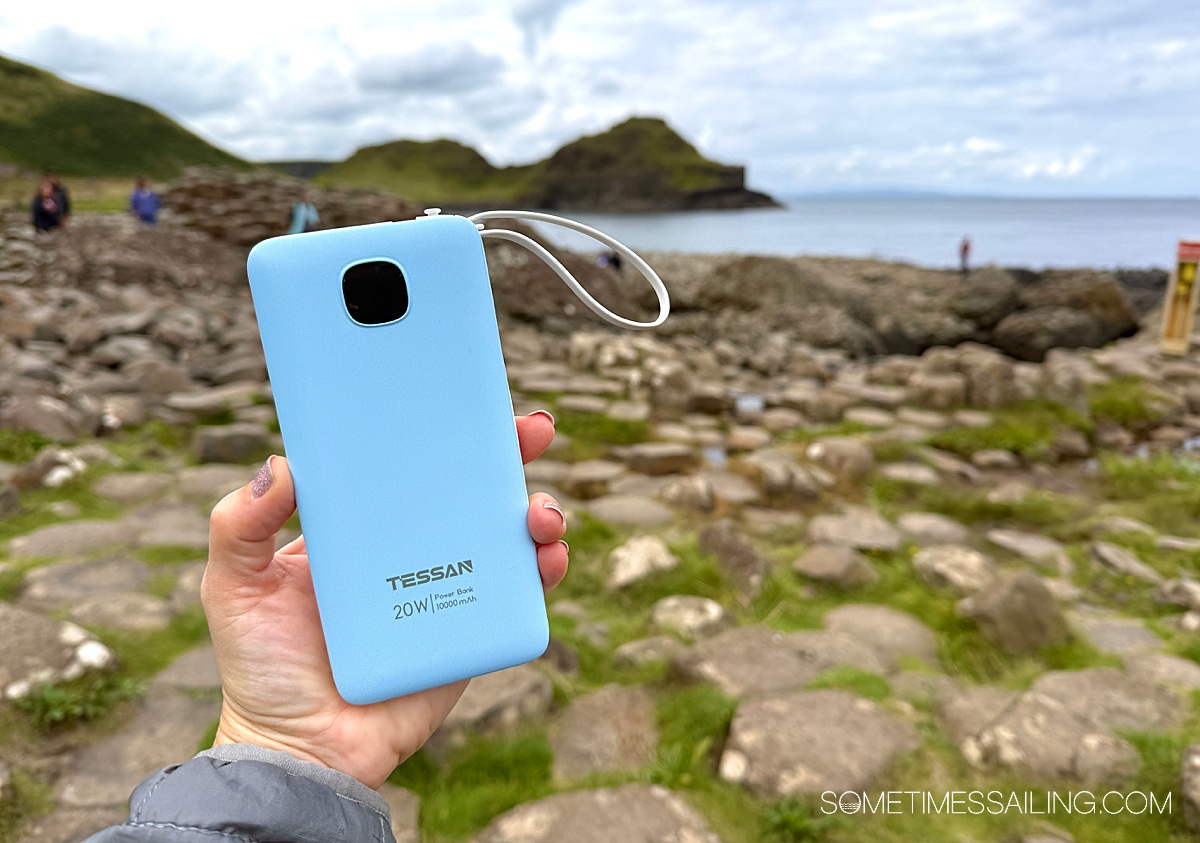 Hand holding a blue rectangular travel battery pack in front of an outdoor ocean-front scene.